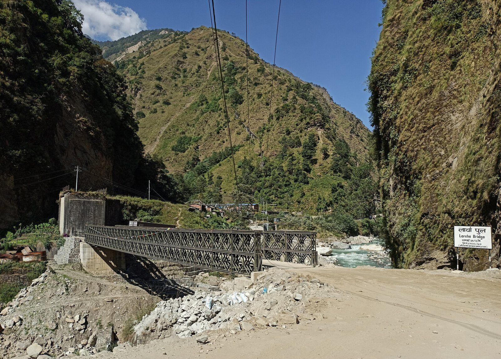 A metal truss bridge spans the fast-flowing Bhotekoshi River in Sindhupalchok District, with piles of construction rubble and rebuilding materials near the riverbank.