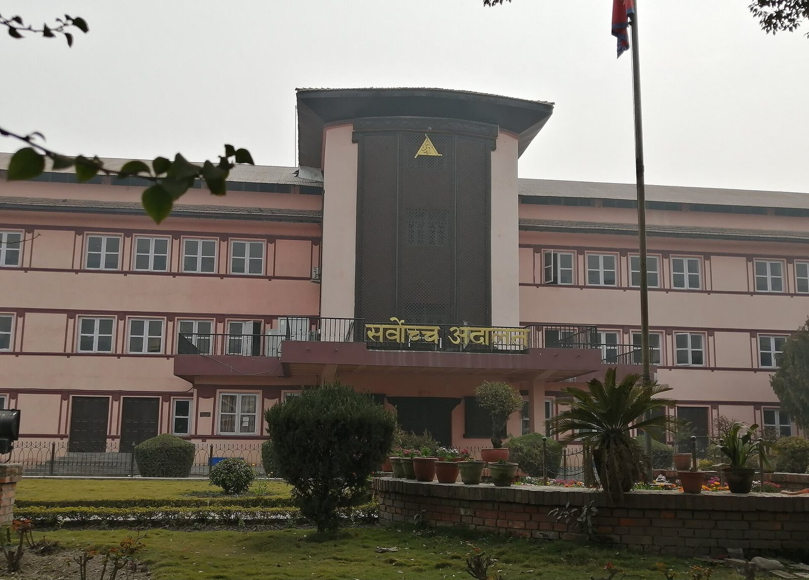 Exterior daytime view of the pink and grey Supreme Court of Nepal building in Kathmandu with its distinctive tower and a Nepali flag out front.