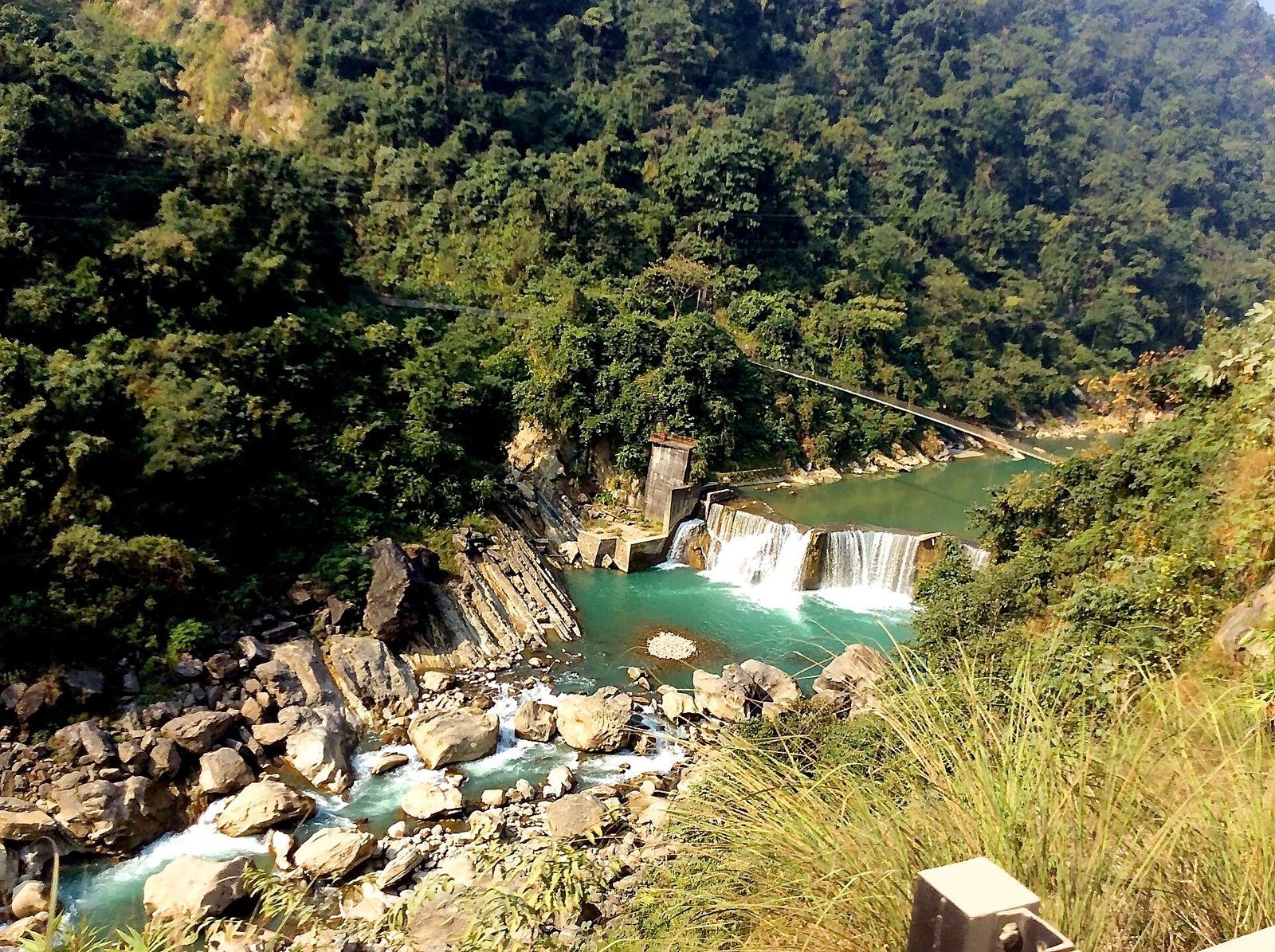 A turquoise river flows past a small dam and rocky banks surrounded by green forest in the Tinau River valley near Butwal, Nepal.