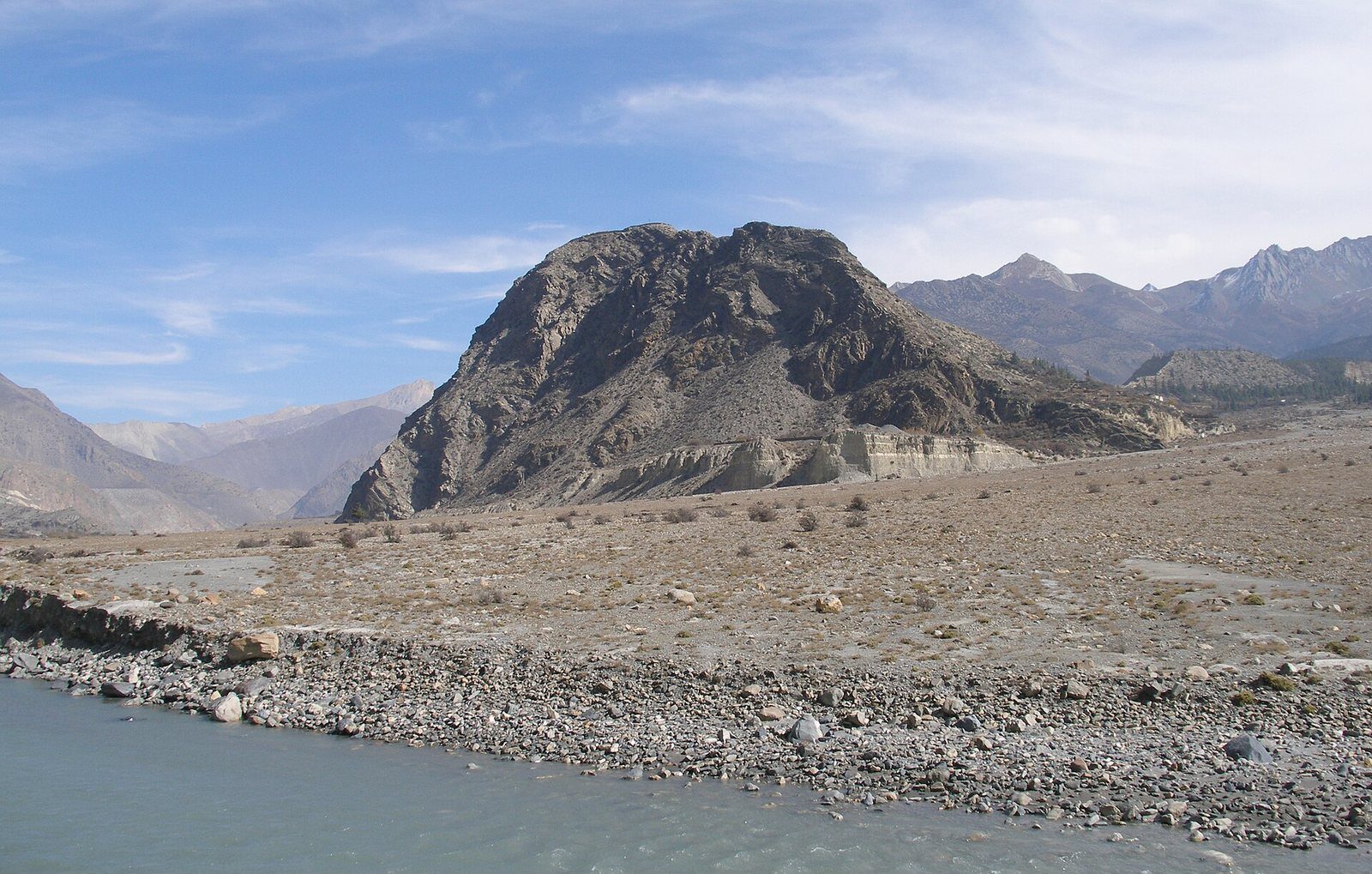 The Kali Gandaki River flows through a broad rocky valley near Jomsom, Nepal, with a large rock outcrop and mountains in the background.
