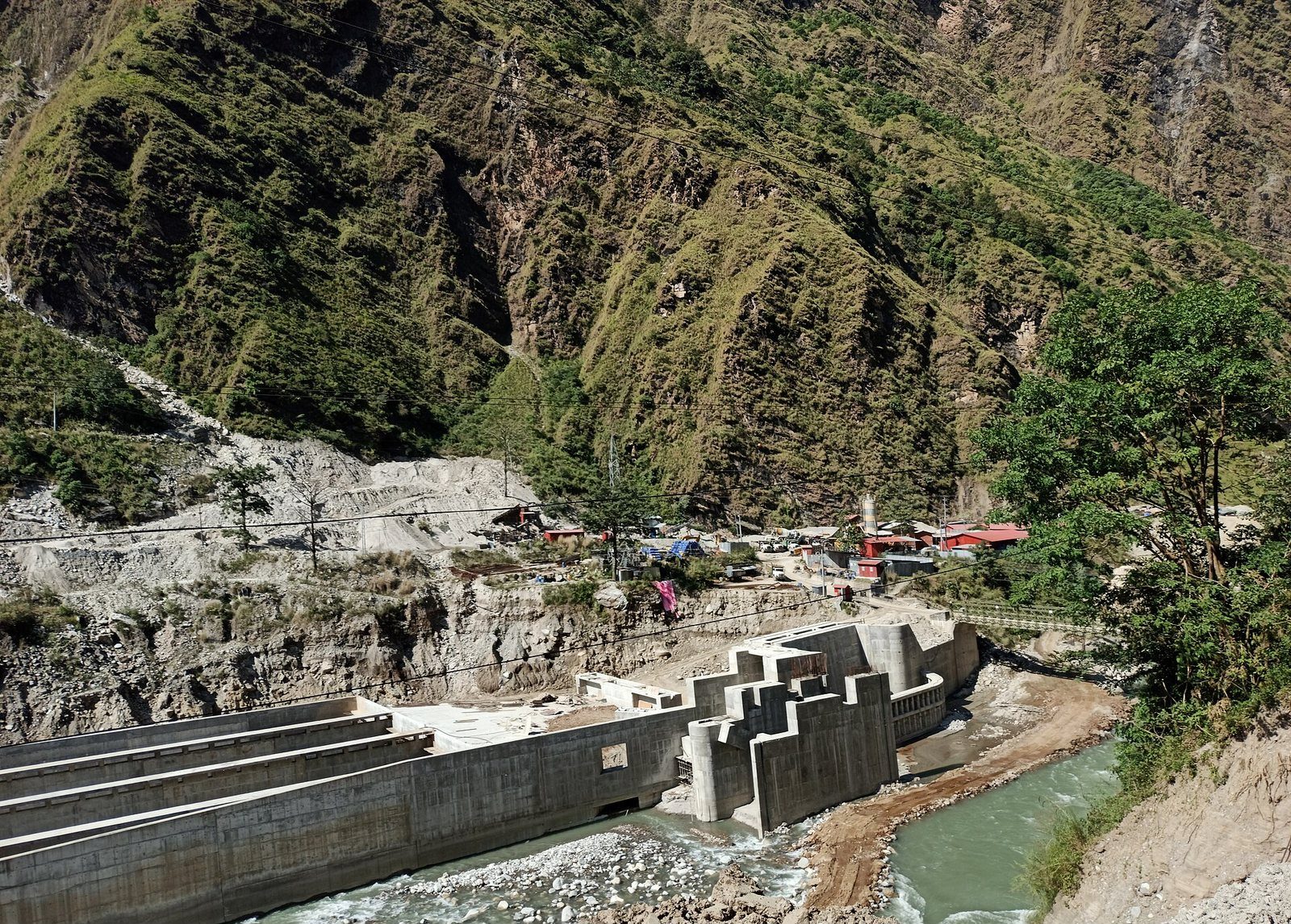 A hydropower project with concrete spillways sits beside the Bhotekoshi River, flanked by steep mountain slopes in Sindhupalchok District, Nepal.