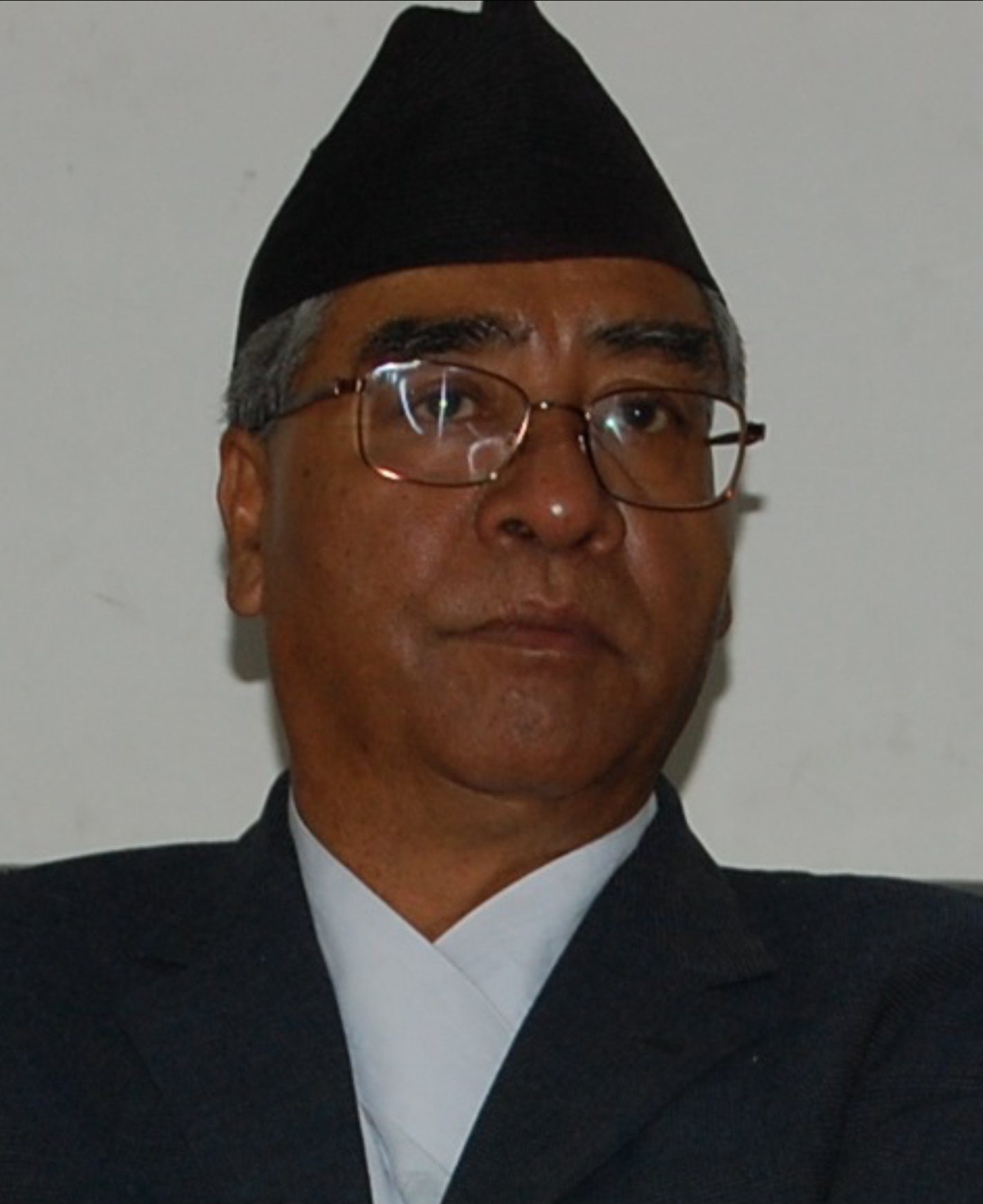 Portrait of Sher Bahadur Deuba wearing a traditional Nepali topi and glasses, looking directly at the camera.