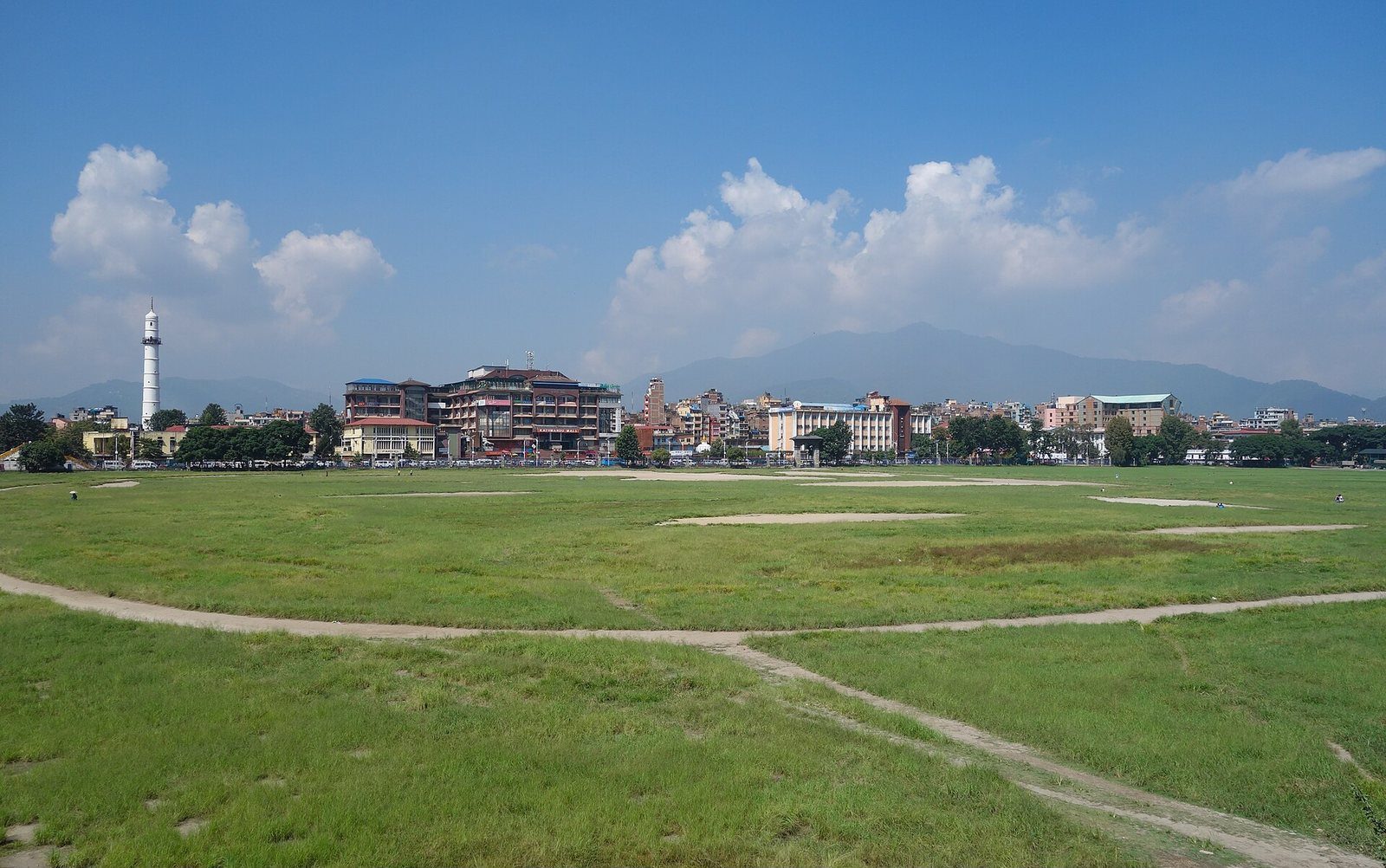 A wide view of the open grassy Tundikhel ground in Kathmandu with city buildings and a tower visible in the background under a blue sky.