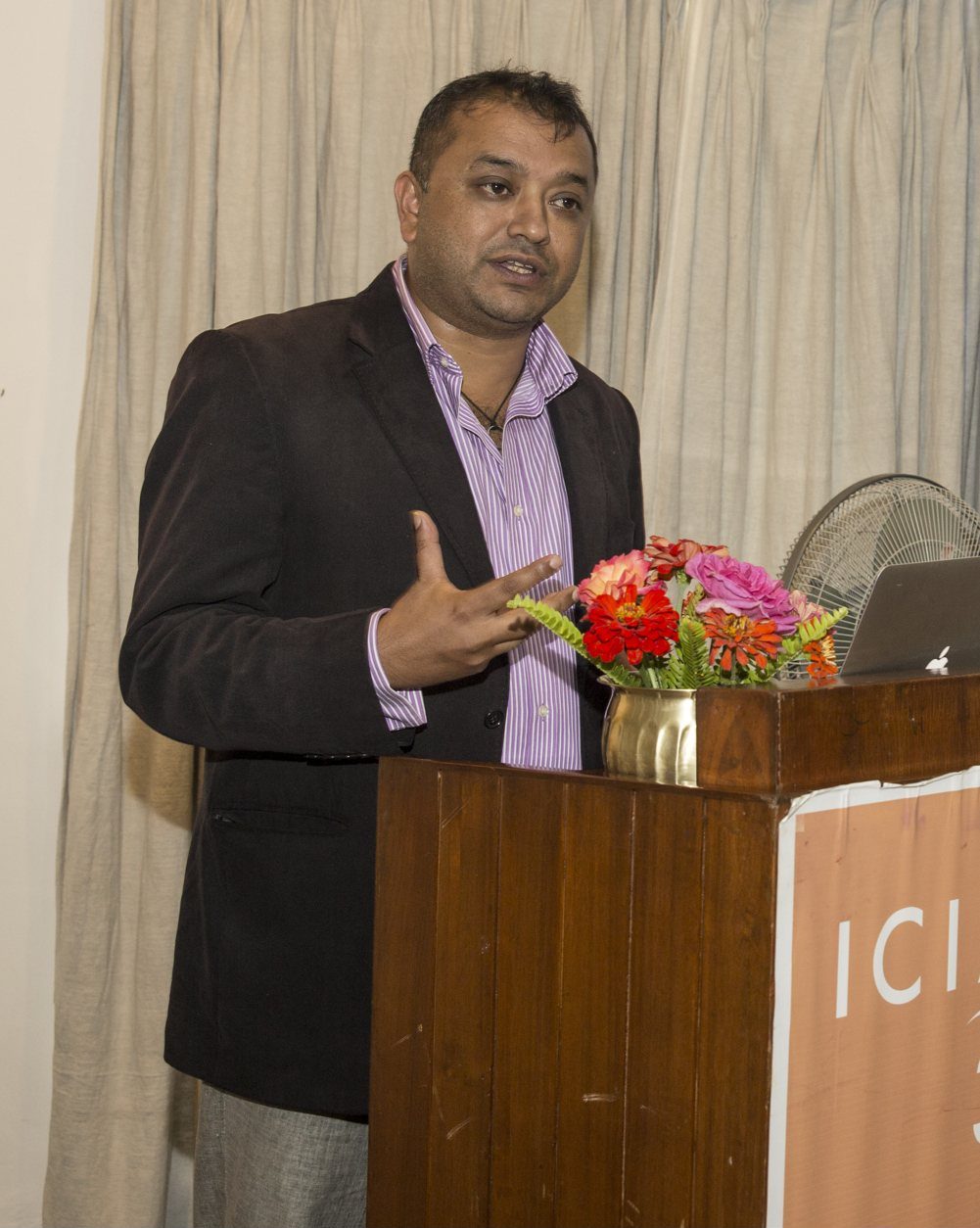 Gagan Thapa, in a dark blazer and striped shirt, gestures while speaking from a wooden podium decorated with flowers.