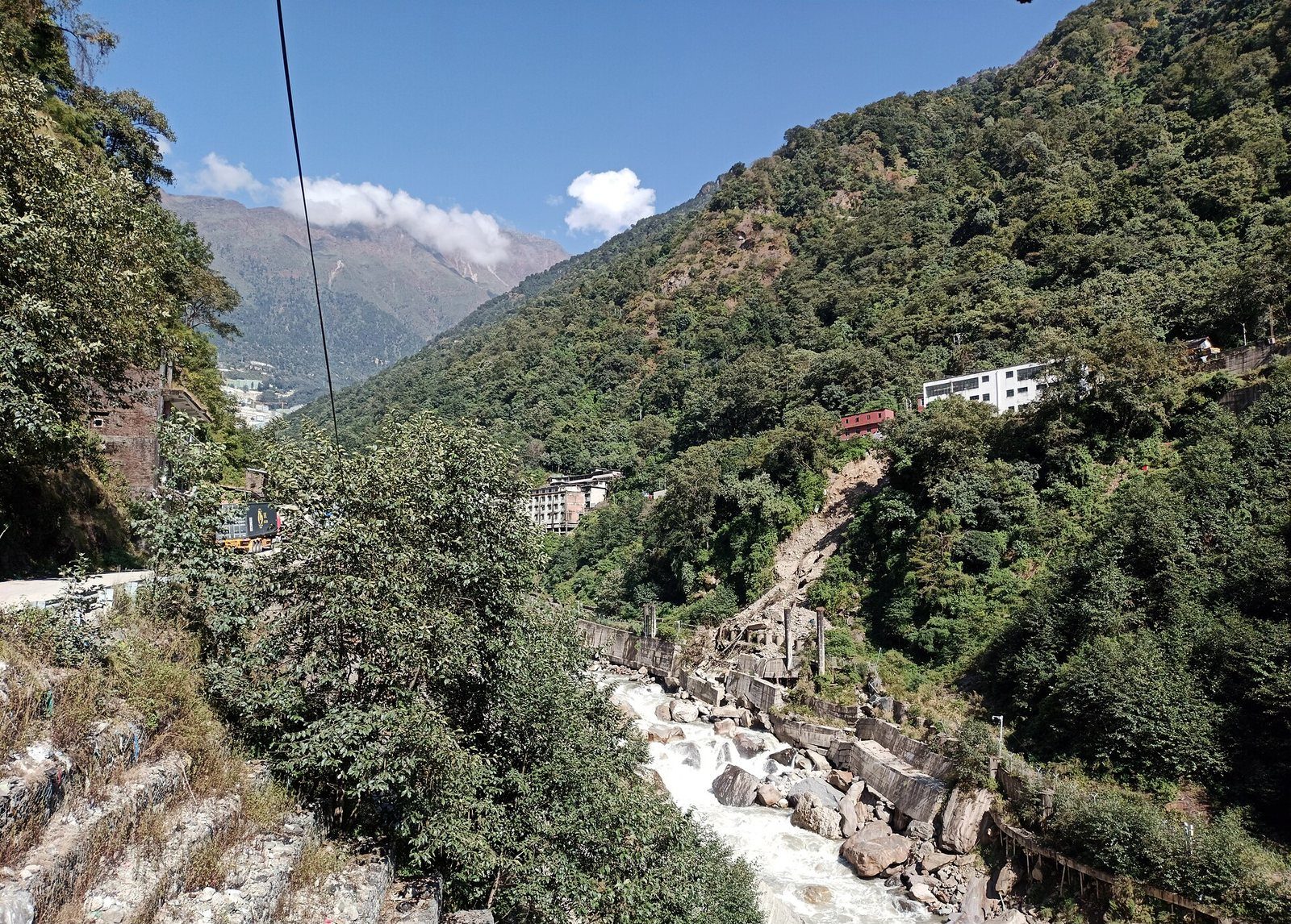 A damaged stretch of the Bhote Koshi River valley in Nepal's hills, with rockslide debris along the banks, a crane working near a collapsed section of road, and forested slopes under a clear sky.