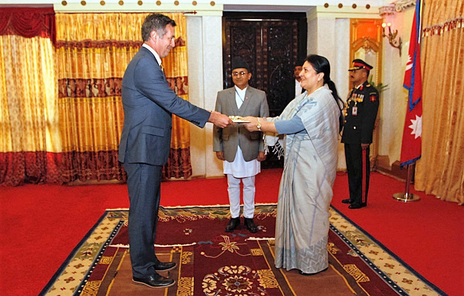 Former President Bidya Devi Bhandari, in a grey sari, receives a document from a foreign ambassador in a formal ceremony hall in Nepal.