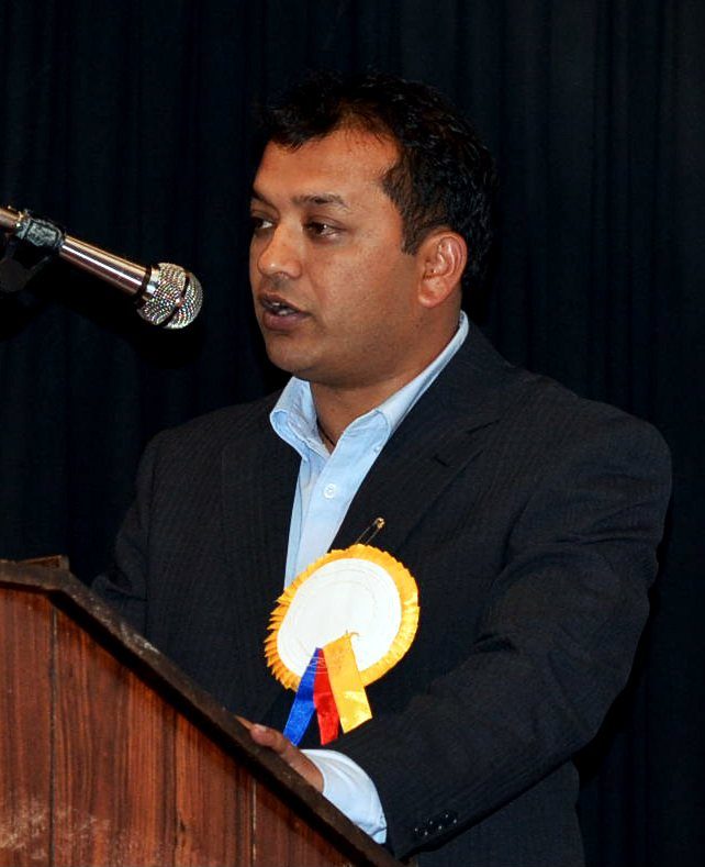 Gagan Thapa, a man in a dark suit, speaks into a microphone at a podium.