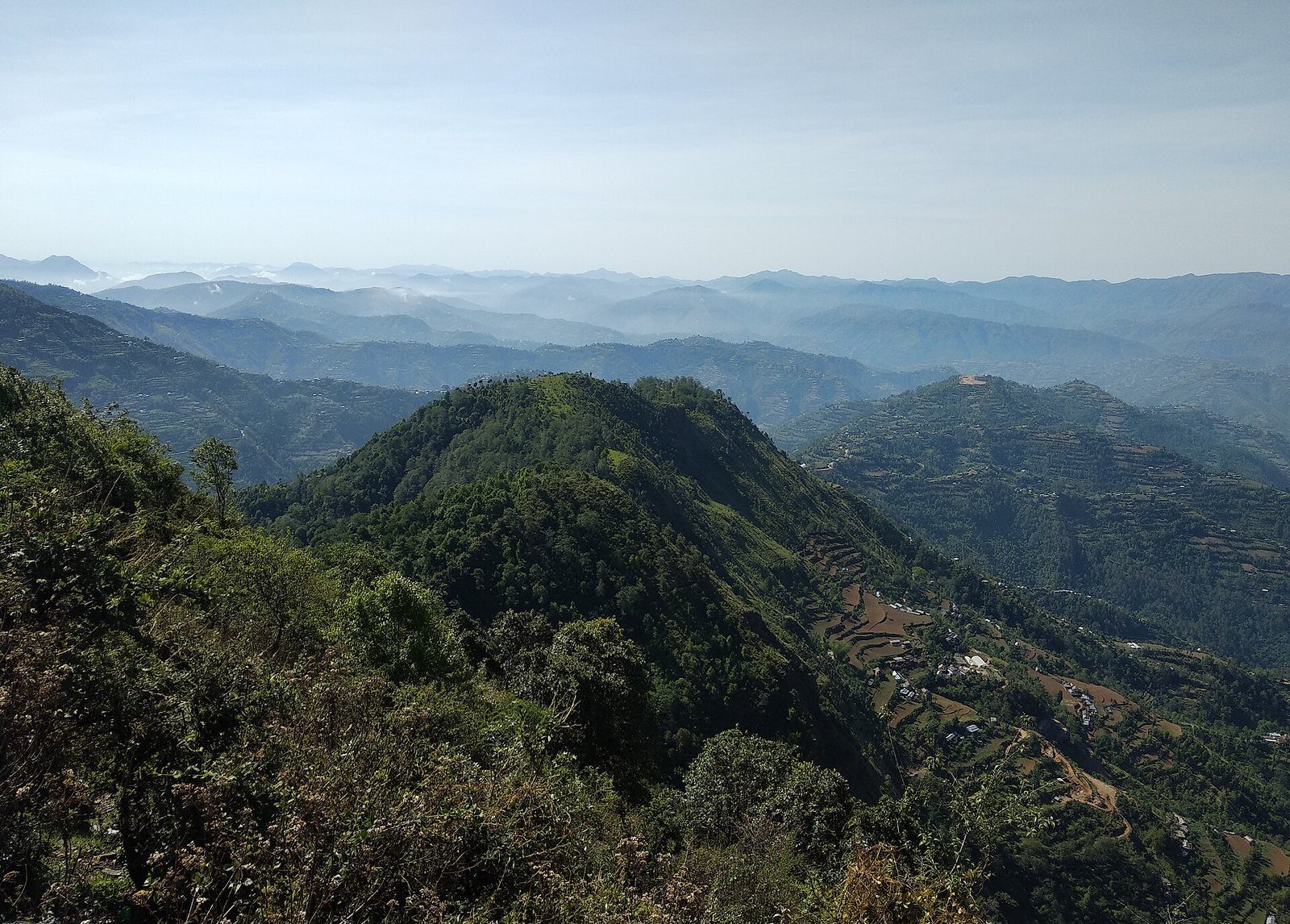 A sweeping view of green terraced hillsides and forested ridges in Gulmi District, Nepal, under a hazy sky.