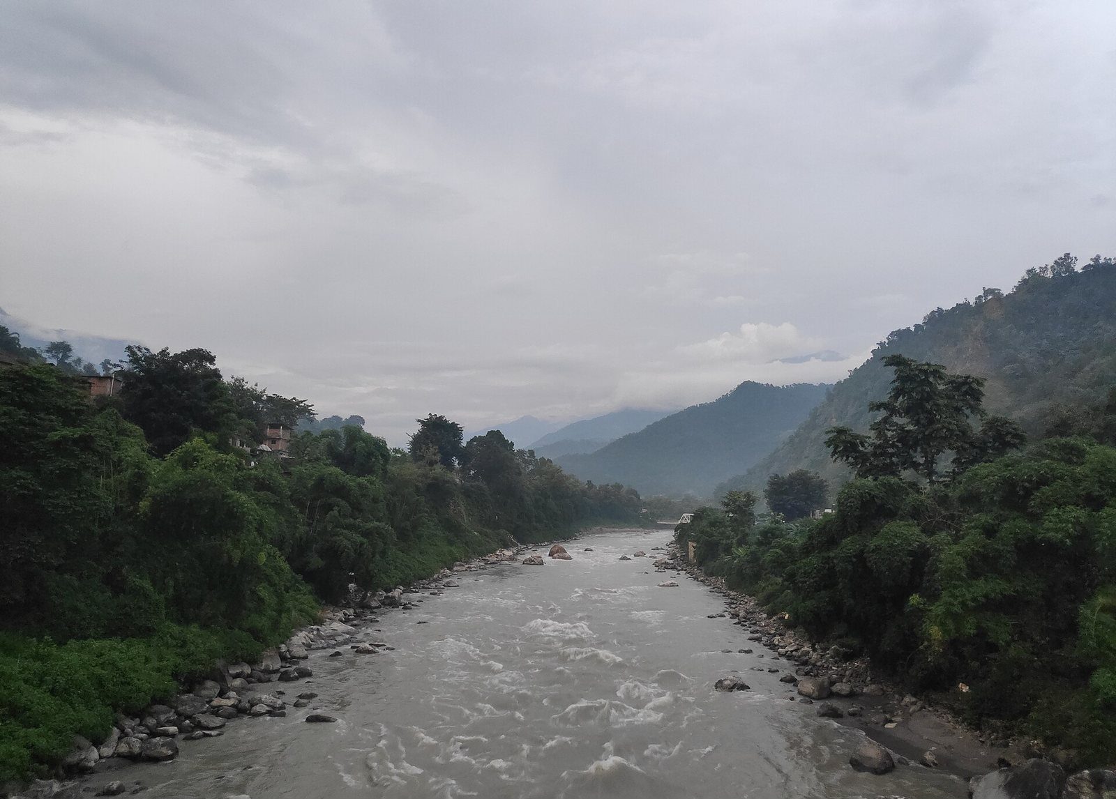 A fast-flowing, murky river winding through a steep green gorge in Nepal's Gorkha district under a cloudy sky.