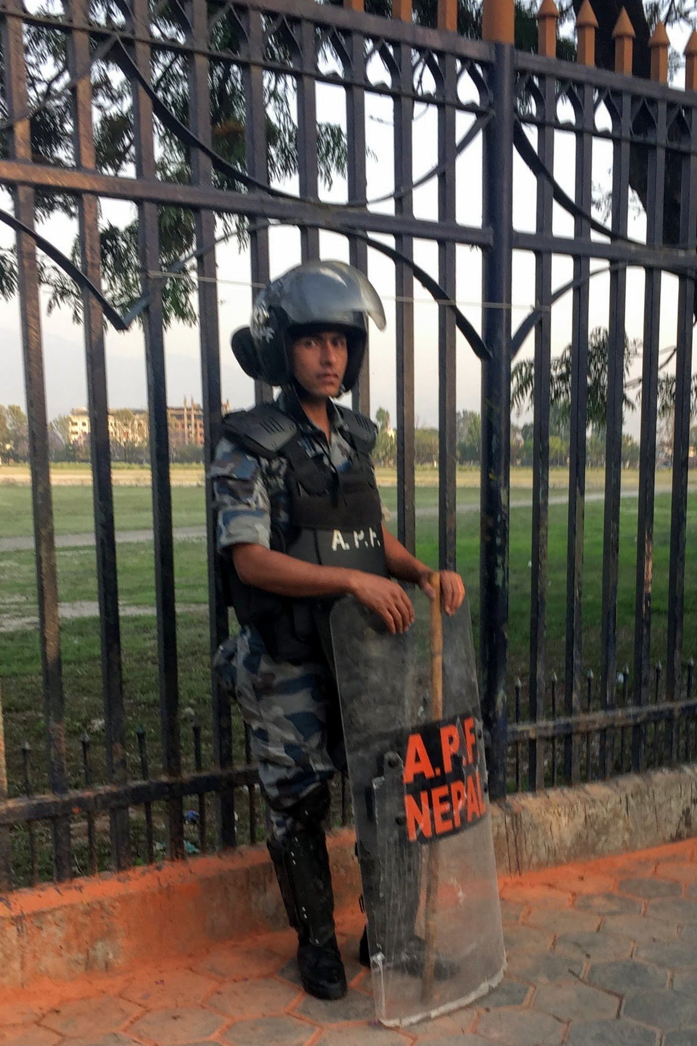 An Armed Police Force of Nepal officer in riot gear with helmet and shield stands beside a metal gate in Kathmandu.