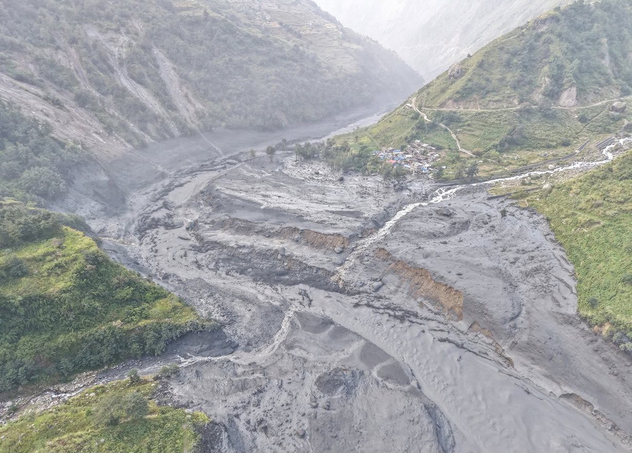 Aerial view of a mud-choked Budhi Gandaki riverbed cutting through a steep Gorkha valley after a landslide dam breach, with a small settlement visible on the bank.