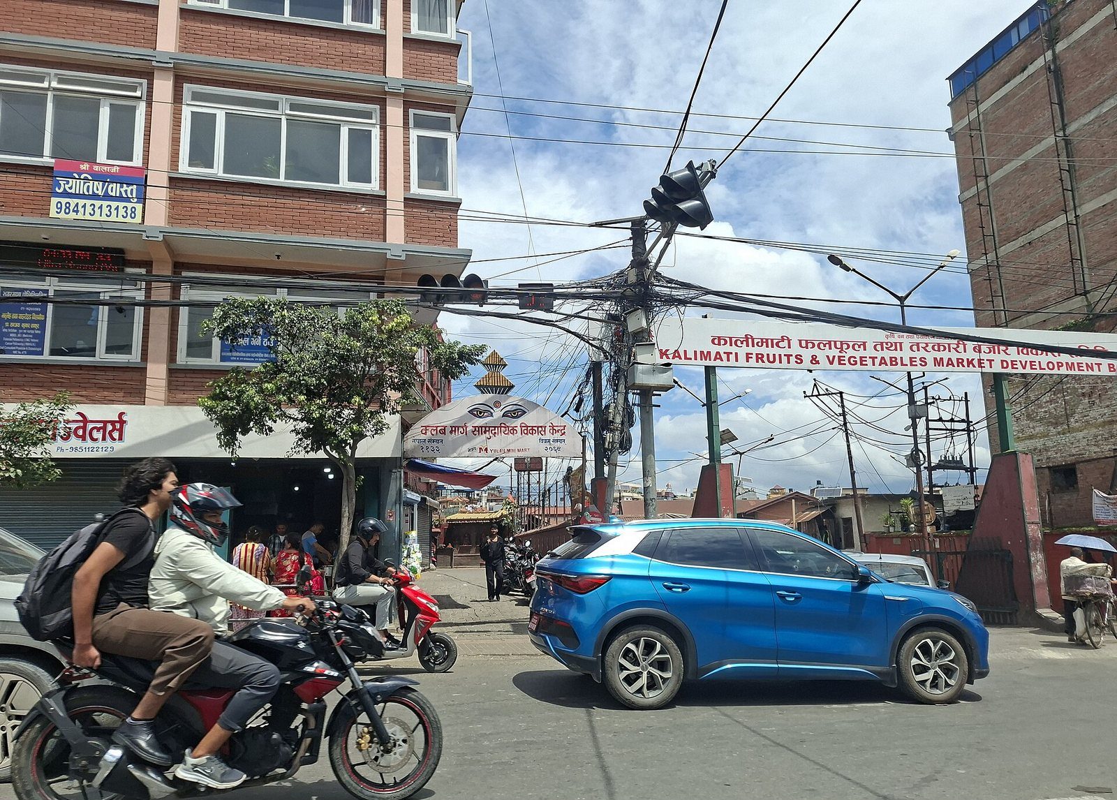 Street view of Kathmandu's Kalimati fruit and vegetable market, with motorbikes and a car passing under the market's signboard.