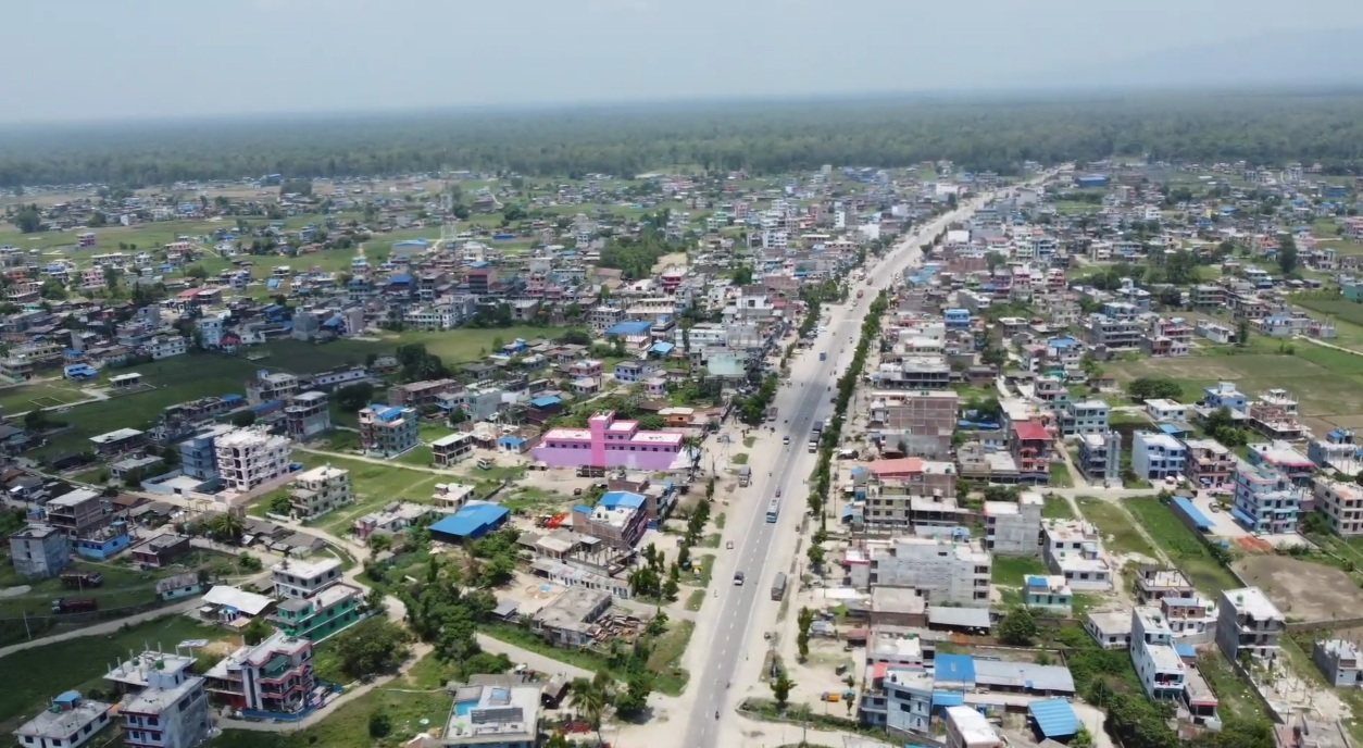 An aerial view of a bustling Tarai town with a straight paved road lined by shops and houses, representing Chandrapur Municipality in Rautahat District.