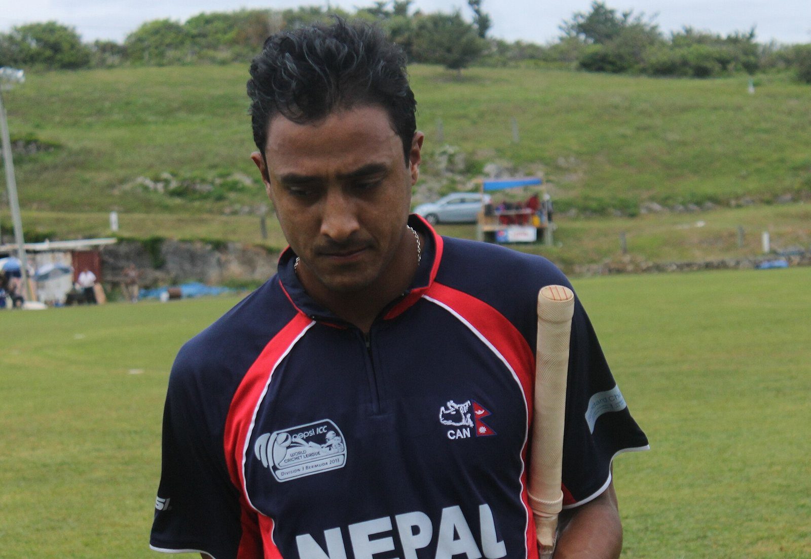 Paras Khadka, wearing a Nepal cricket team jersey, stands on a grassy pitch holding a cricket bat.