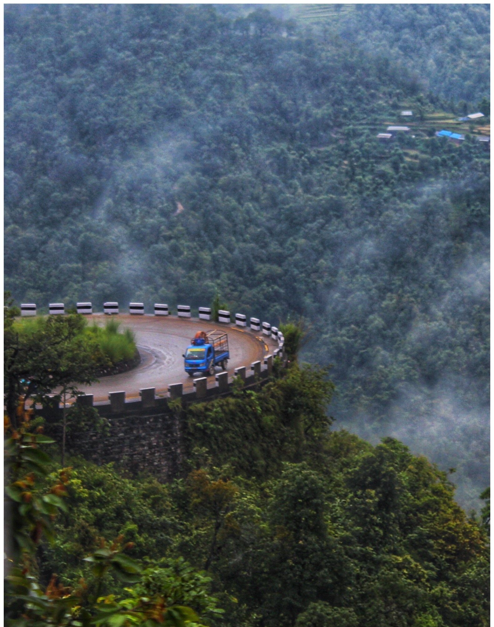 A winding section of the BP Highway cutting through forested hills in Sindhuli district, Nepal, with a truck navigating a sharp curve.