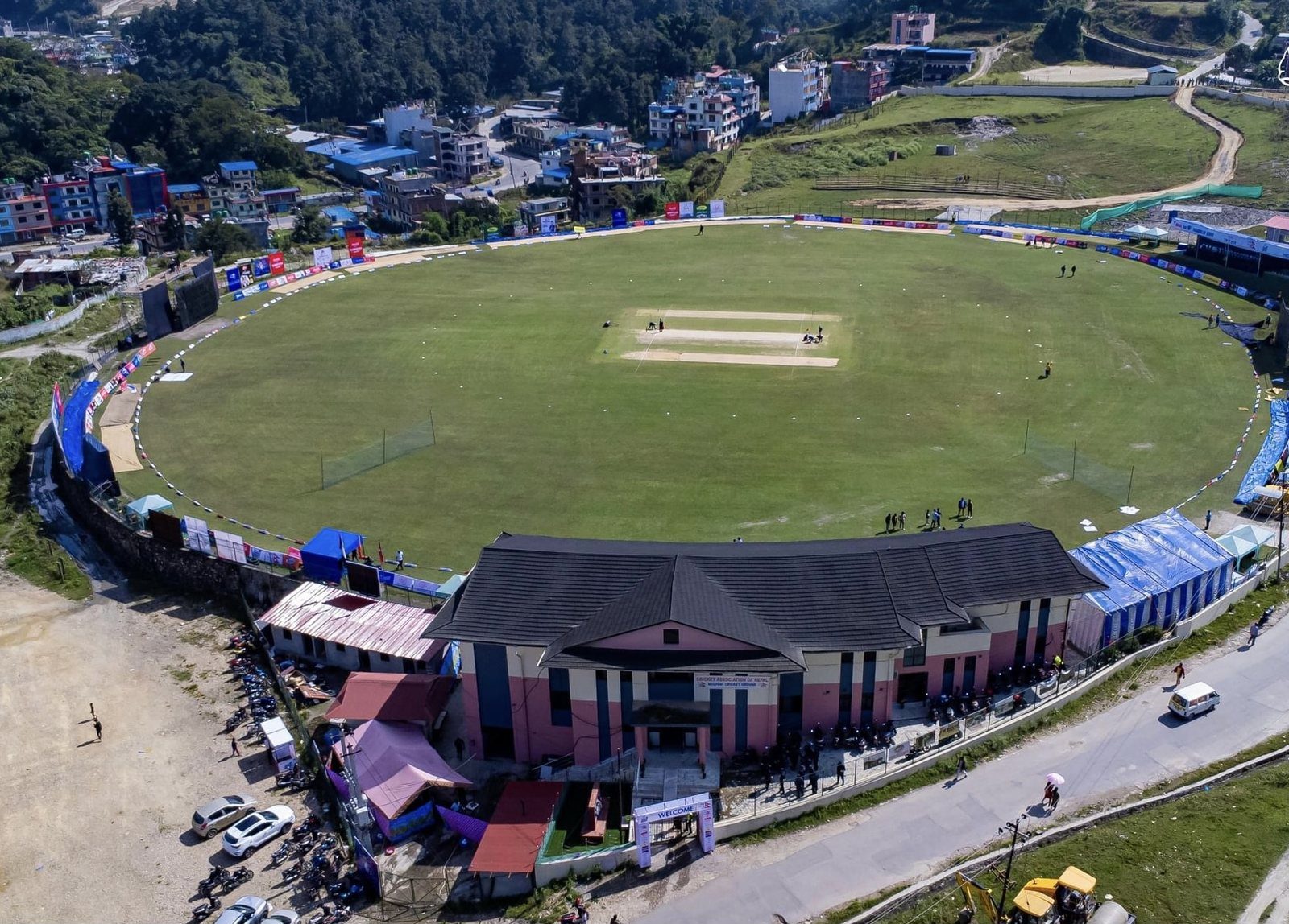 An aerial view of the Mulpani Cricket Ground in Kathmandu, showing the oval pitch, surrounding stands and a hillside town beyond.