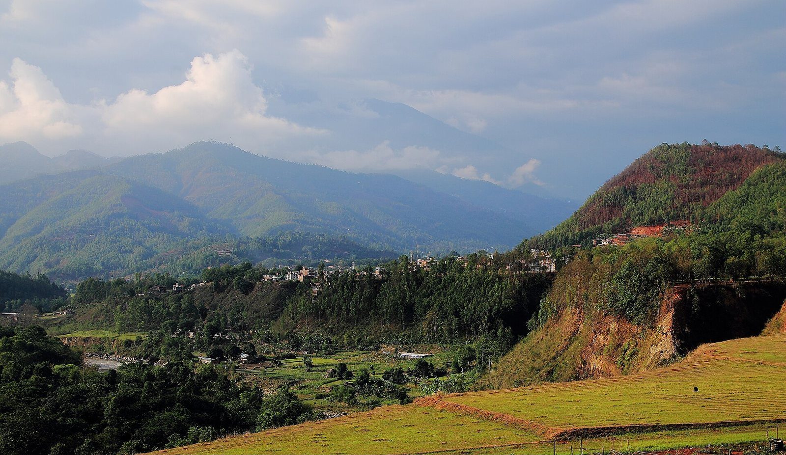A wide green valley landscape in Nuwakot District, Nepal, with hills and scattered settlements under a hazy sky.