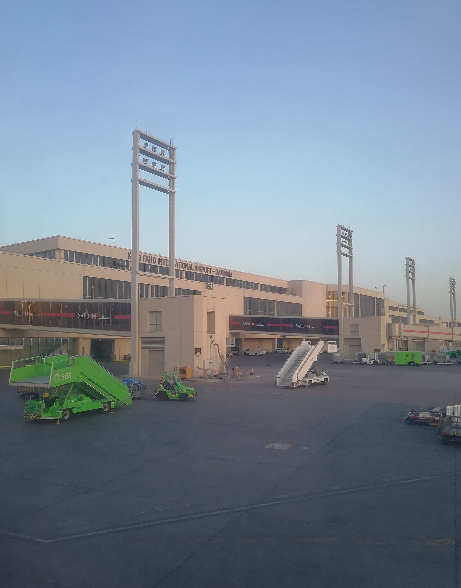 Exterior view of King Fahd International Airport in Dammam, Saudi Arabia, showing the terminal building, light towers and tarmac with ground vehicles under a clear sky.