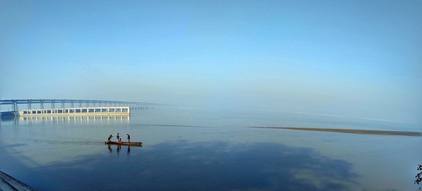 A wide view of the Koshi River with a barrage bridge in the background and men boating on the water.