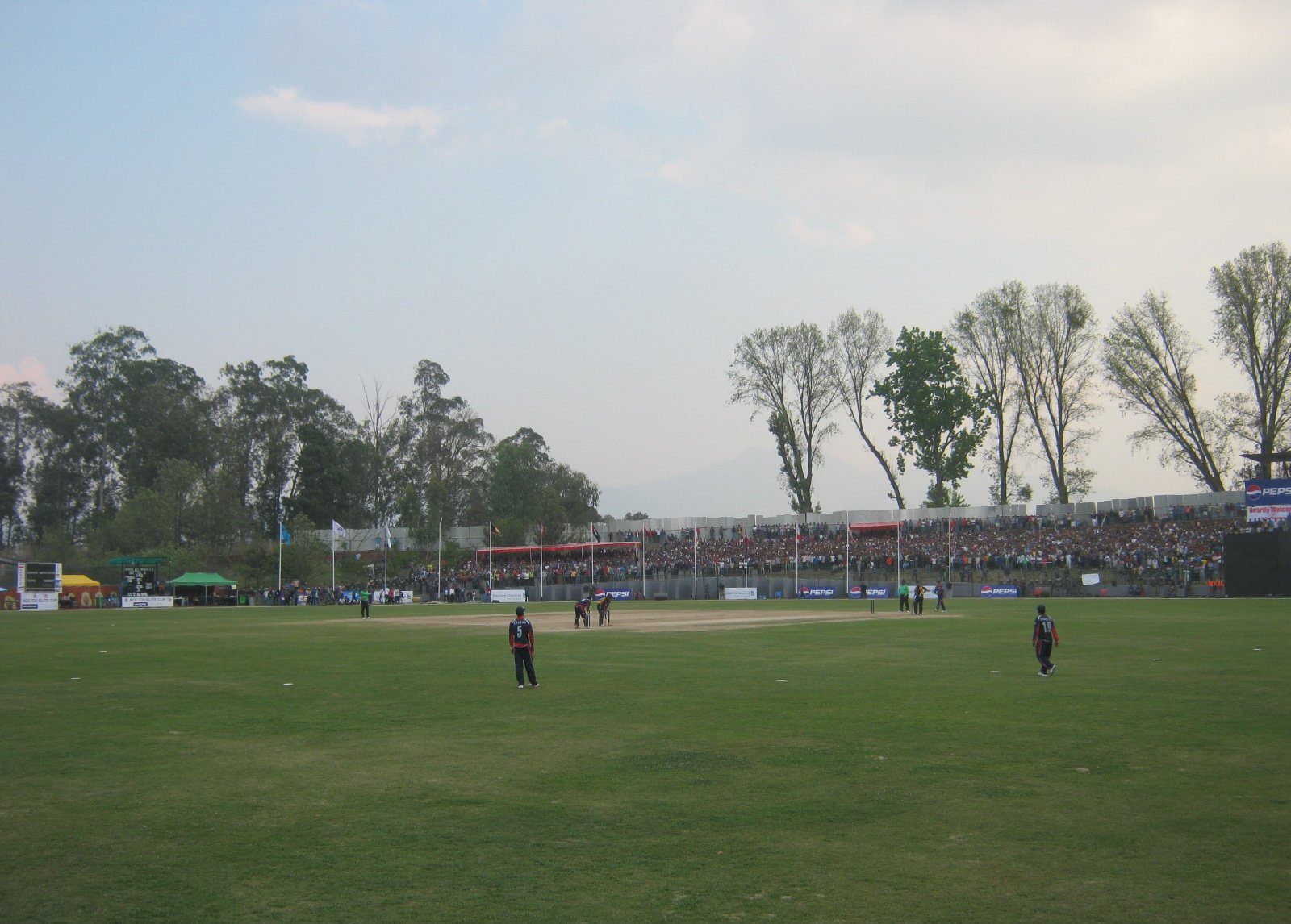 A cricket match in progress on the field at Tribhuvan University International Cricket Ground, with players positioned around the pitch and spectators filling the stands in the background.