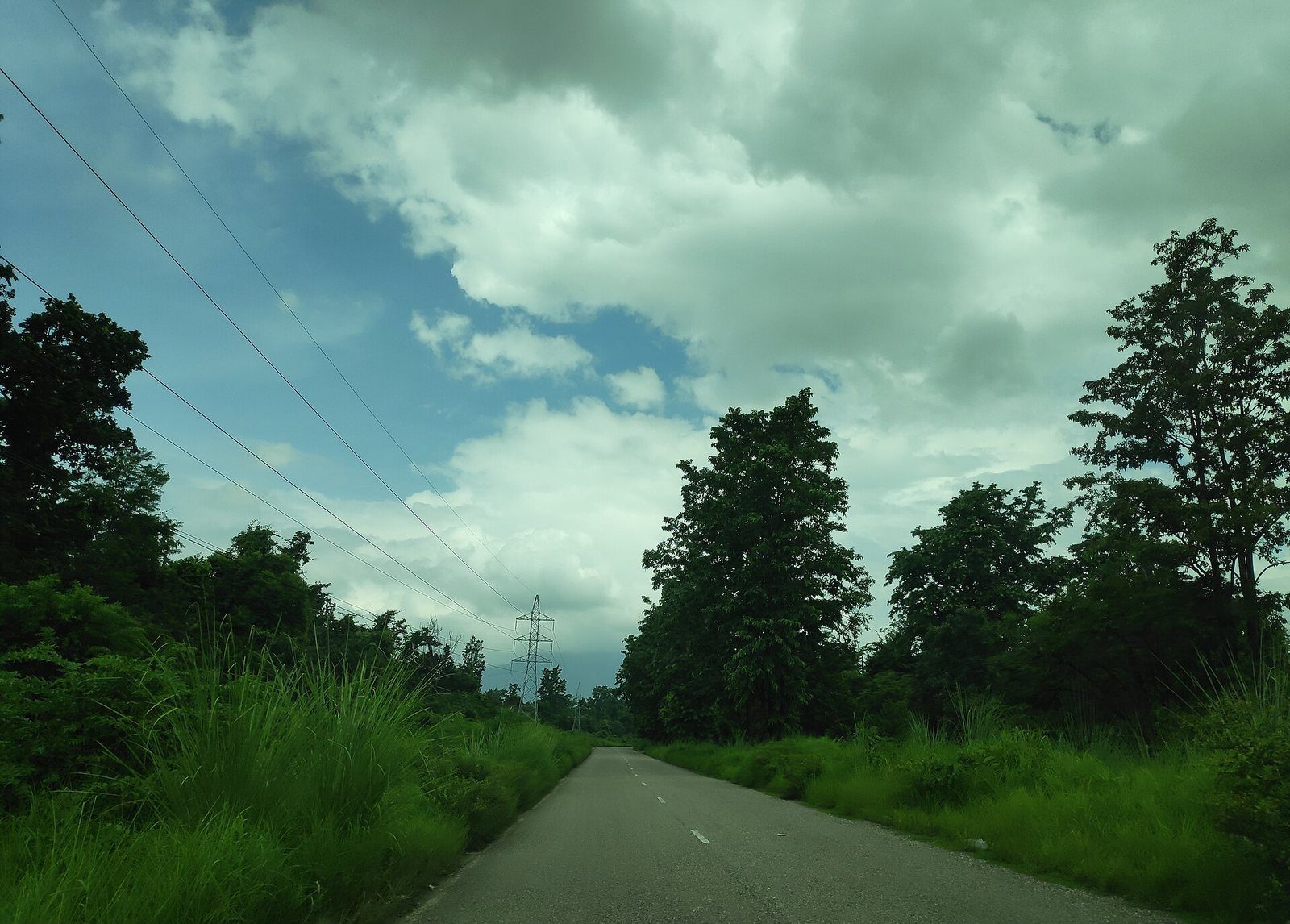An empty paved highway running through a forested, cloudy landscape in Nepal, flanked by power lines and greenery.