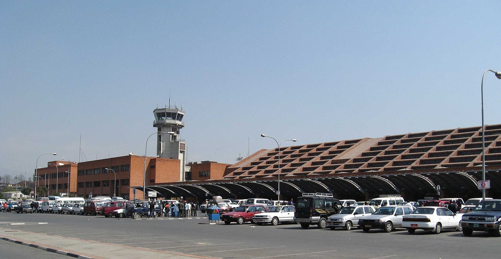The terminal building and control tower of Tribhuvan International Airport in Kathmandu, with vehicles parked outside.