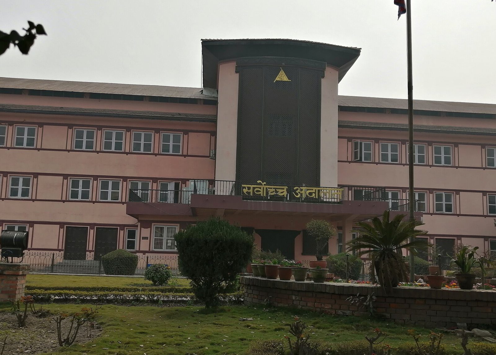 The pink and white Supreme Court of Nepal building in Kathmandu, with its Nepali signage and flagpole, seen from the courtyard.