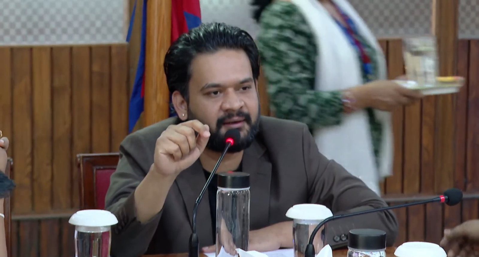 Balen Shah, bearded and in a brown blazer, speaks into a microphone at a formal table with a Nepal flag in the background.